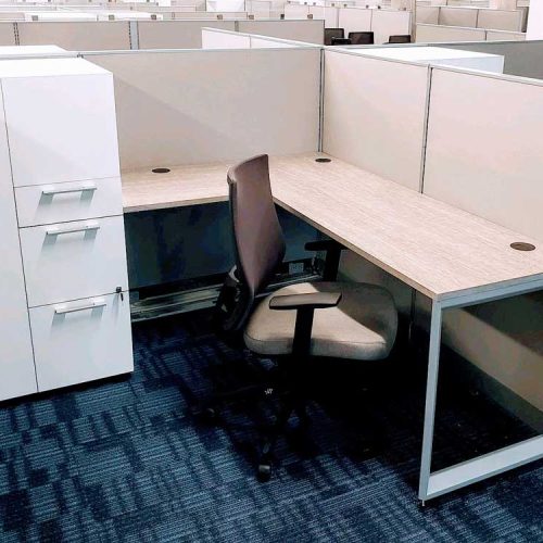 An office cubicle featuring a corner desk, ergonomic chair, and white storage cabinets in a spacious, organized workspace.