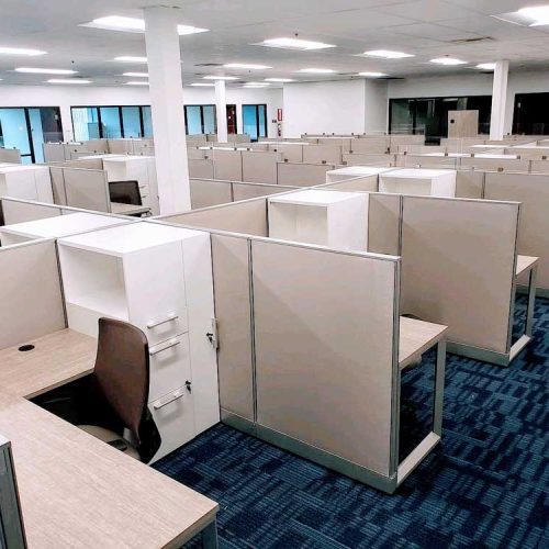 An empty office space featuring numerous cubicles with beige partitions, desks, and chairs, all on a blue patterned carpet.
