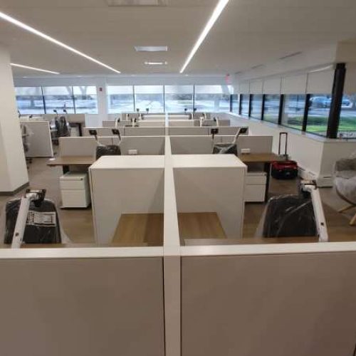 An empty office space with cubicles, desks, and large windows, illuminated by overhead lights, waiting for setup.