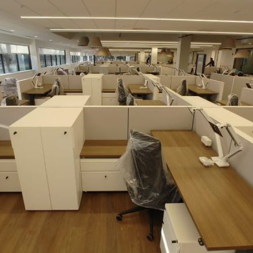 A modern, spacious office with multiple cubicles featuring wooden desks, plastic-covered chairs, and large windows.