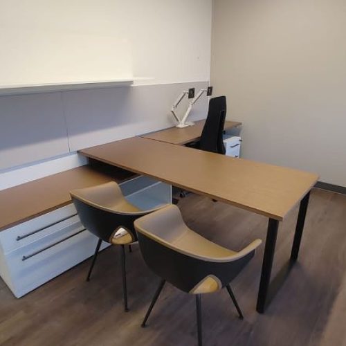 A modern office setup featuring a long desk, two chairs, storage drawers, and wall-mounted shelves in a minimalistic room.
