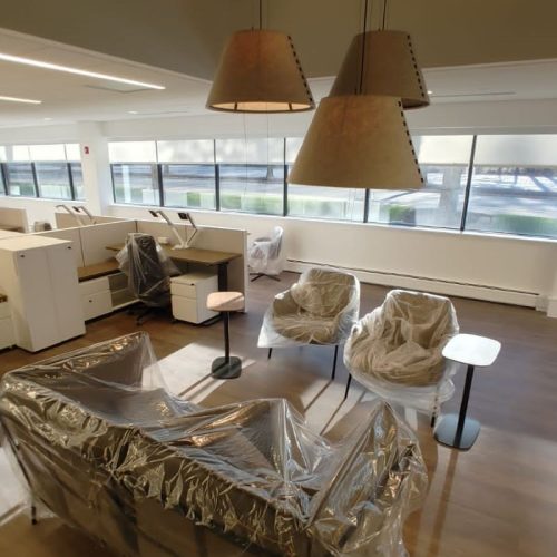 An office space with a couch and chairs covered in plastic, two lamps hanging above, and large windows letting in light.