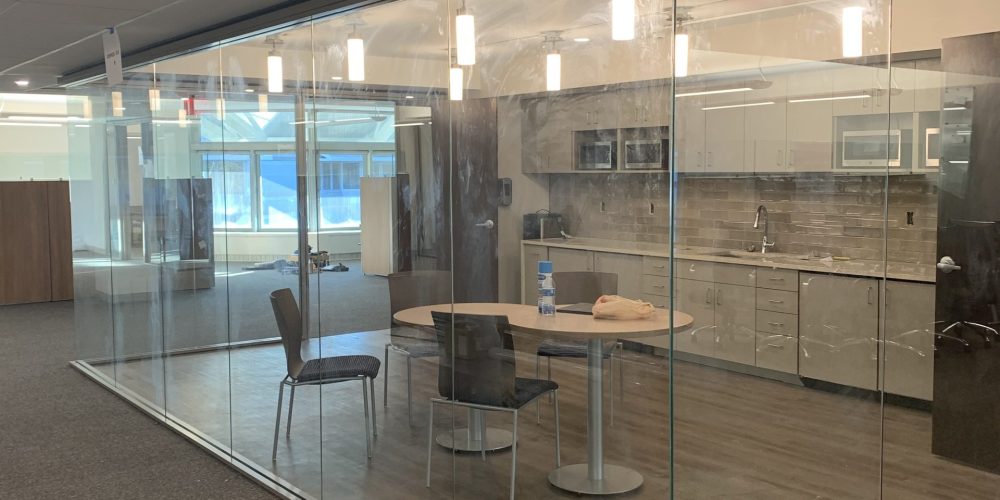 A modern glass meeting room with a round table and chairs, illuminated by pendant lights, situated in an office space.