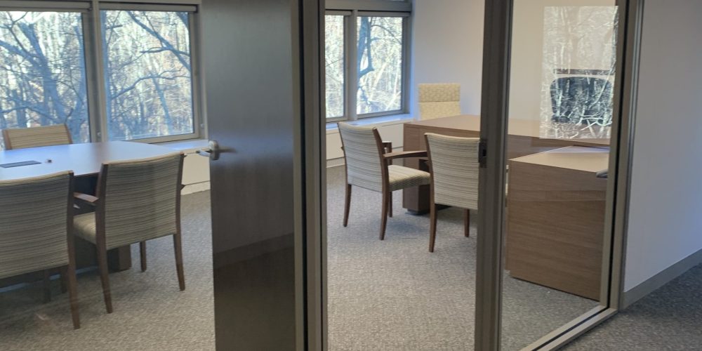 A modern conference room with glass walls, featuring a large table and wooden chairs, illuminated by natural light.