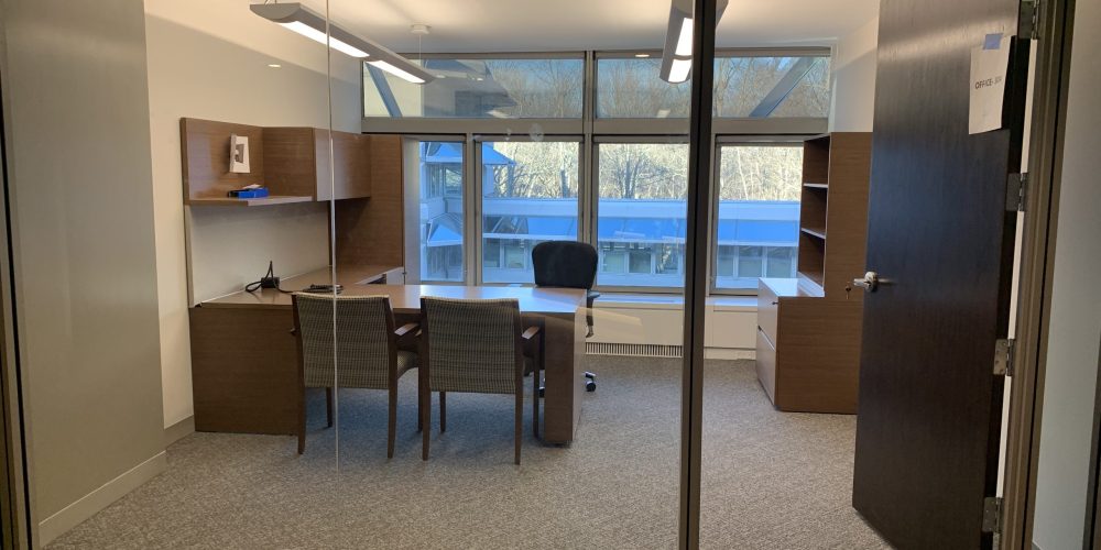 A modern office with a glass wall, featuring a desk, two chairs, bookshelves, and large windows offering natural light.