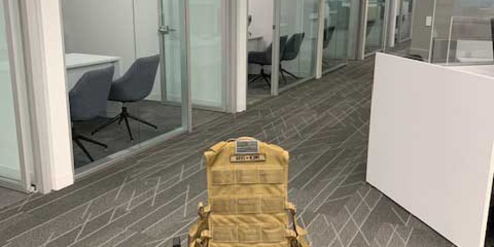 A wheeled cart with a tan backpack stands in an empty, modern office corridor, featuring glass walls and minimalist decor.