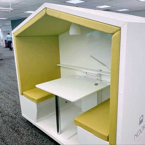 A modern workspace nook featuring a white frame with yellow cushions, a small table, and overhead lighting, designed for privacy.