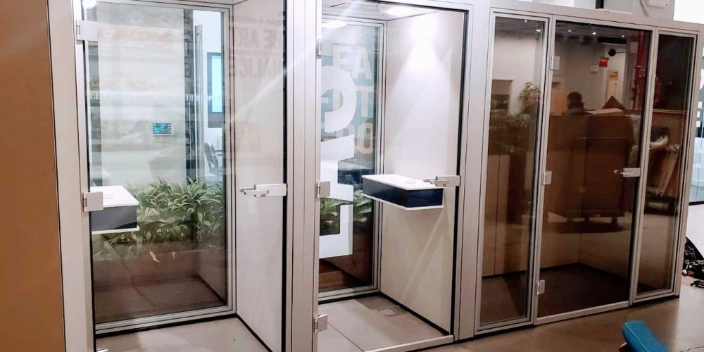 A modern, glass-enclosed workspace with four booths, featuring minimalist design and greenery visible inside.