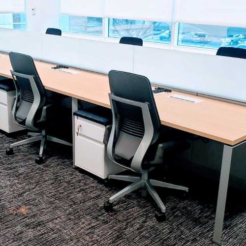 An office space featuring desks and chairs arranged in front of large windows, allowing natural light to illuminate the area.