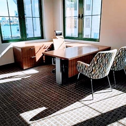 A modern office with large windows, a wooden desk, ergonomic chair, and two patterned chairs on a plush carpet.